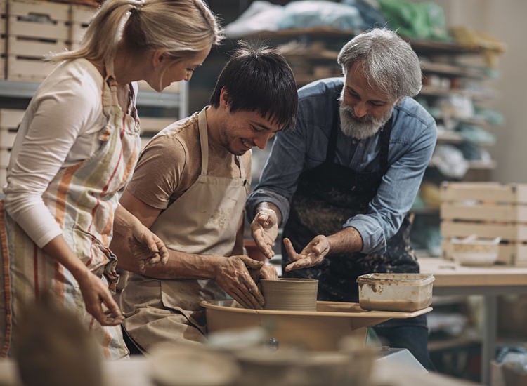 Tre mennesker er samlet omkring en drejeskive og former ler i et atelier